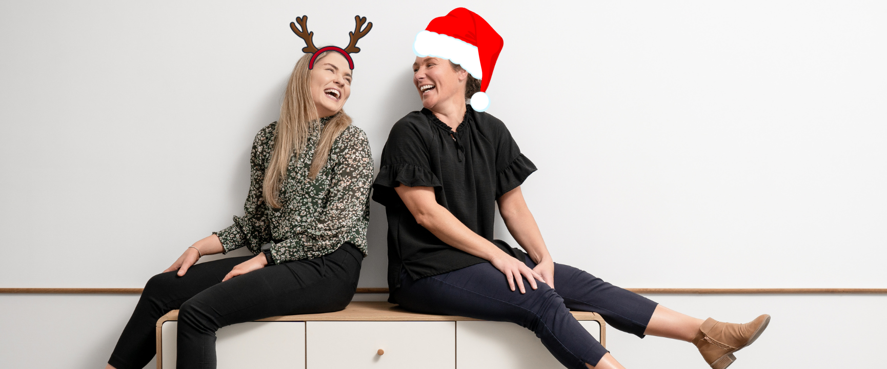 Sarah and Beck sit back to back on against a white wall. They are laughing and looking at each other. Sarah has cartoon reindeer antlers on her head. Beck has a cartoon santa hat, both of which have been edited on. 