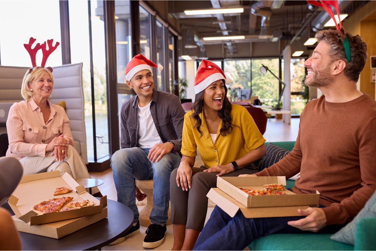 Four colleagues sitting around a table eating pizza with Christmas hats on.
